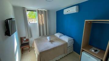 A hotel room with a bed, a desk, a chair, a TV, and an air conditioning unit.