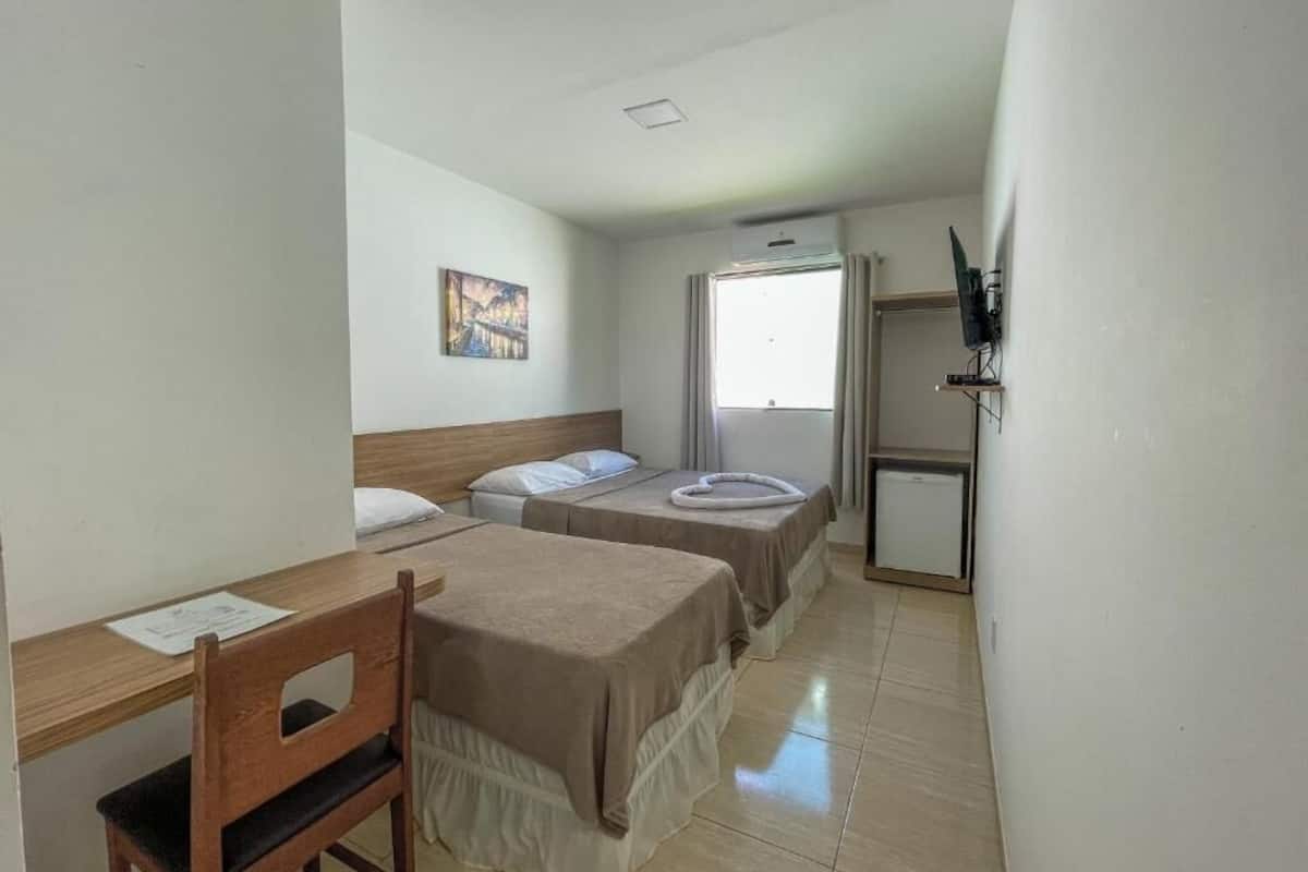 A hotel room with two beds, a desk, a chair, a small fridge, and a window with curtains.