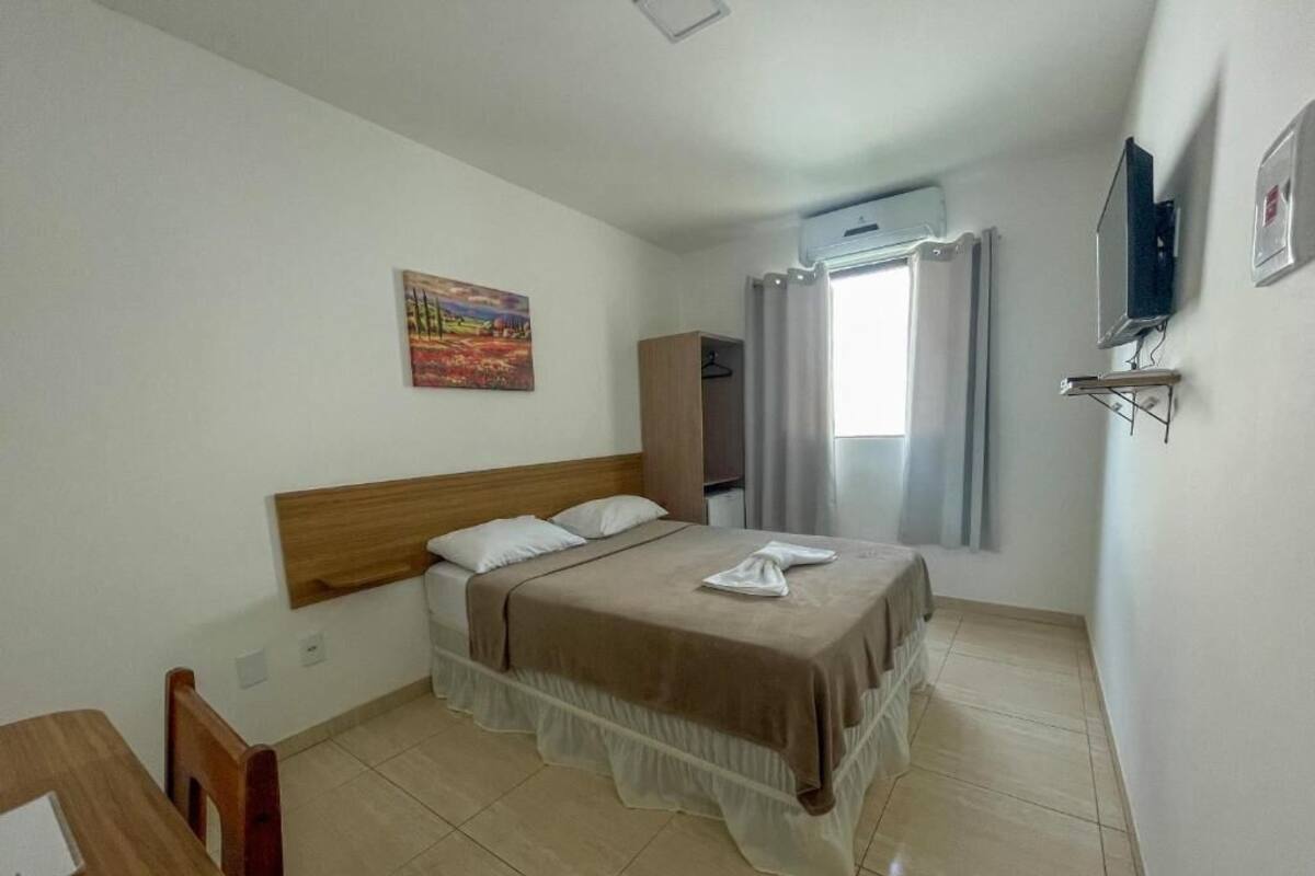 A hotel room with a bed, a wooden headboard, a painting on the wall, an air conditioning unit, and a window with curtains.