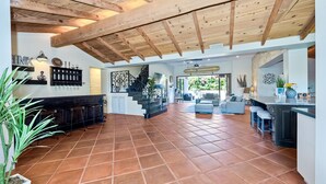 Ampla sala de estar com piso ladrilhado, vigas de madeira no teto, uma escada e um espaço de cozinha com banquetas.