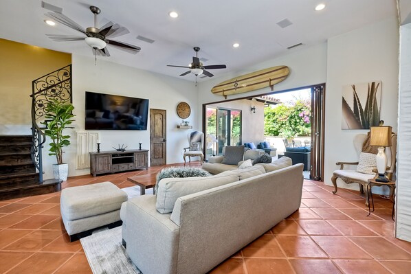 A spacious living room with a large sofa, a ceiling fan, a surfboard decoration, and a television. - Walk To The Beach | Outdoor Dining | Spacious (Carlsbad)