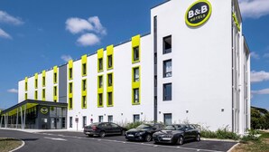 A modern B&B hotel with a white facade and yellow accents, featuring a large parking area with several cars.