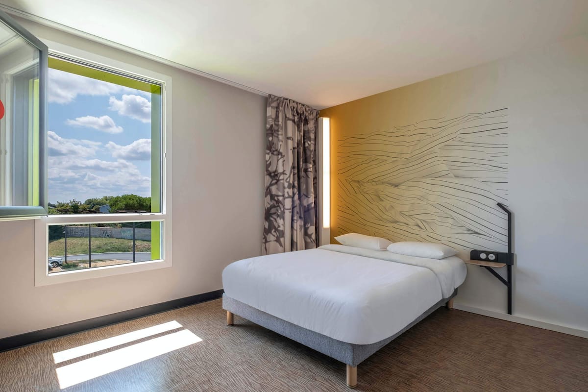 A modern hotel room with a large bed, a view of the outdoors through a window, and a unique wall art piece.