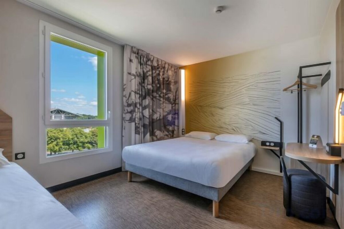 A hotel room with a bed, a desk, a chair, a window with a view, and a wall-mounted lamp.