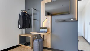 A modern hotel room with a flat-screen TV mounted on the wall, a laptop on a wooden desk, a chair, a coat rack, and a shoe storage unit.