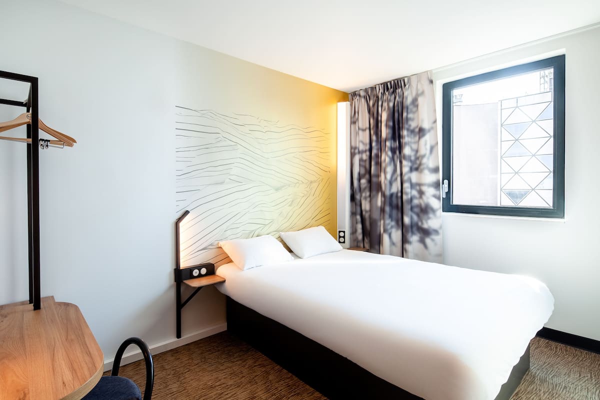 A modern hotel room with a neatly made bed, a wooden chair, a small table with a lamp, and a window with curtains.