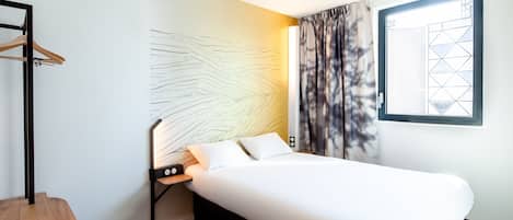 A modern hotel room with a neatly made bed, a wooden chair, a small table with a lamp, and a window with curtains.