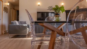 A modern living room with a glass dining table, clear chairs, a grey sofa, and a TV mounted on the wall. - Stormfront Guesthouse-Downtown, Hot Tub, Arcade Games (Tofino)