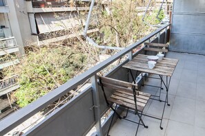 A balcony with a wooden table and chairs, a metal railing, and a view of a building with balconies and trees. - Charming 2 Bedroom apt next to Panormou (Athens)