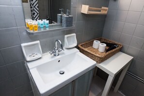 Ein modernes Badezimmer mit einem weißen Waschbecken, einem chromfarbenen Wasserhahn und einem Spiegel darüber. Auf einem Regal stehen Pflegeprodukte und ein Korbwäscher mit Handtüchern.