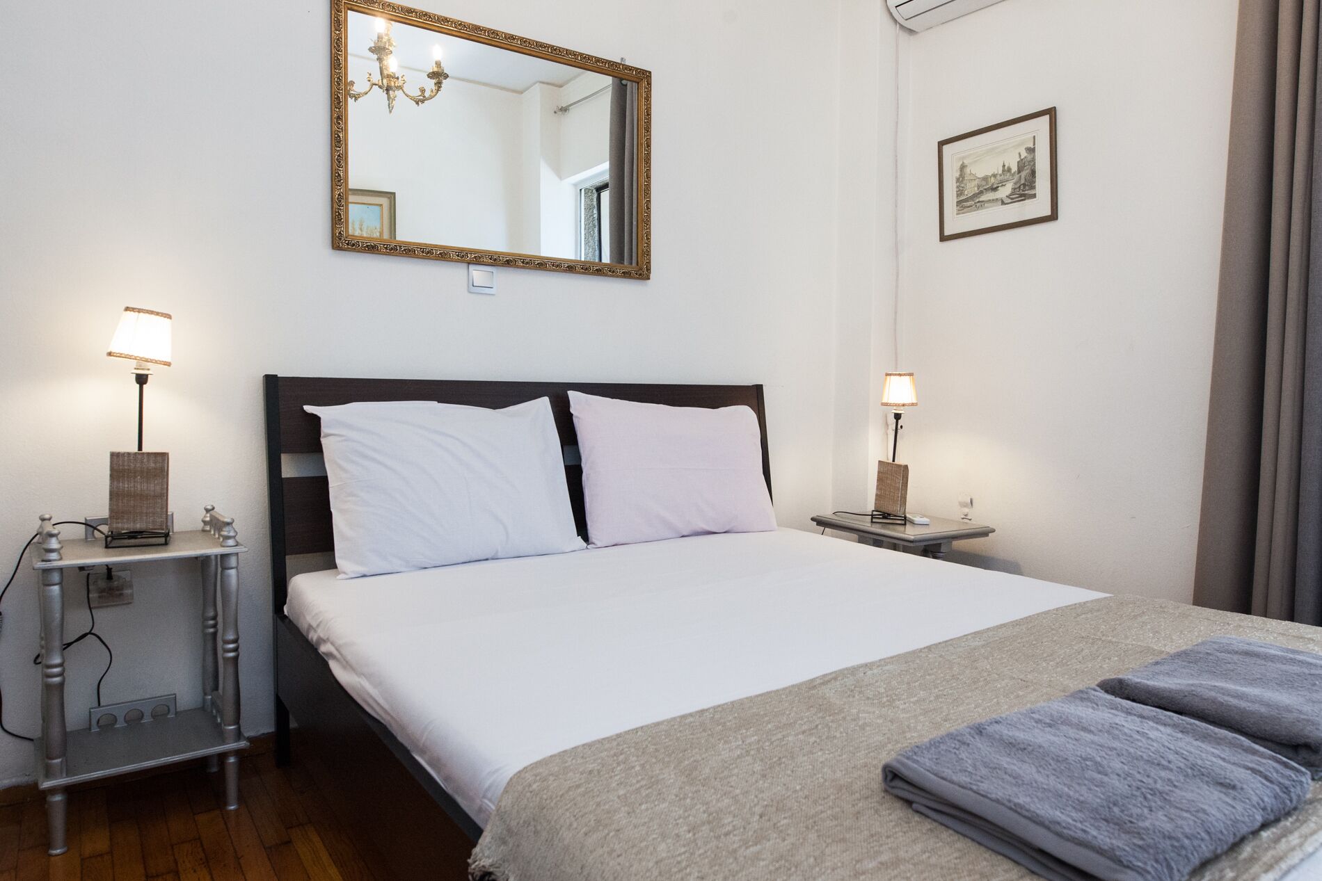 A neatly arranged hotel room with a bed, bedside lamps, a bedside table, a mirror, and a framed picture on the wall.