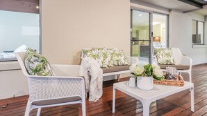 Een moderne zithoek met wit rieten meubilair, een bank met palmbladkussens en een bijzettafel met decoratieve objecten.