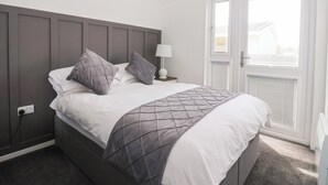 A neatly made bed with a grey headboard, white bedding, and a grey quilted throw. There is a bedside table with a lamp and a power outlet. - Bonny Bay (Bridlington)