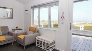 A modern, well-lit room with a grey sofa, a small white table, and a bench. There are two windows with blinds, one of which has a sign reading "I am staying in chalet Number 428". - Bonny Bay (Bridlington)
