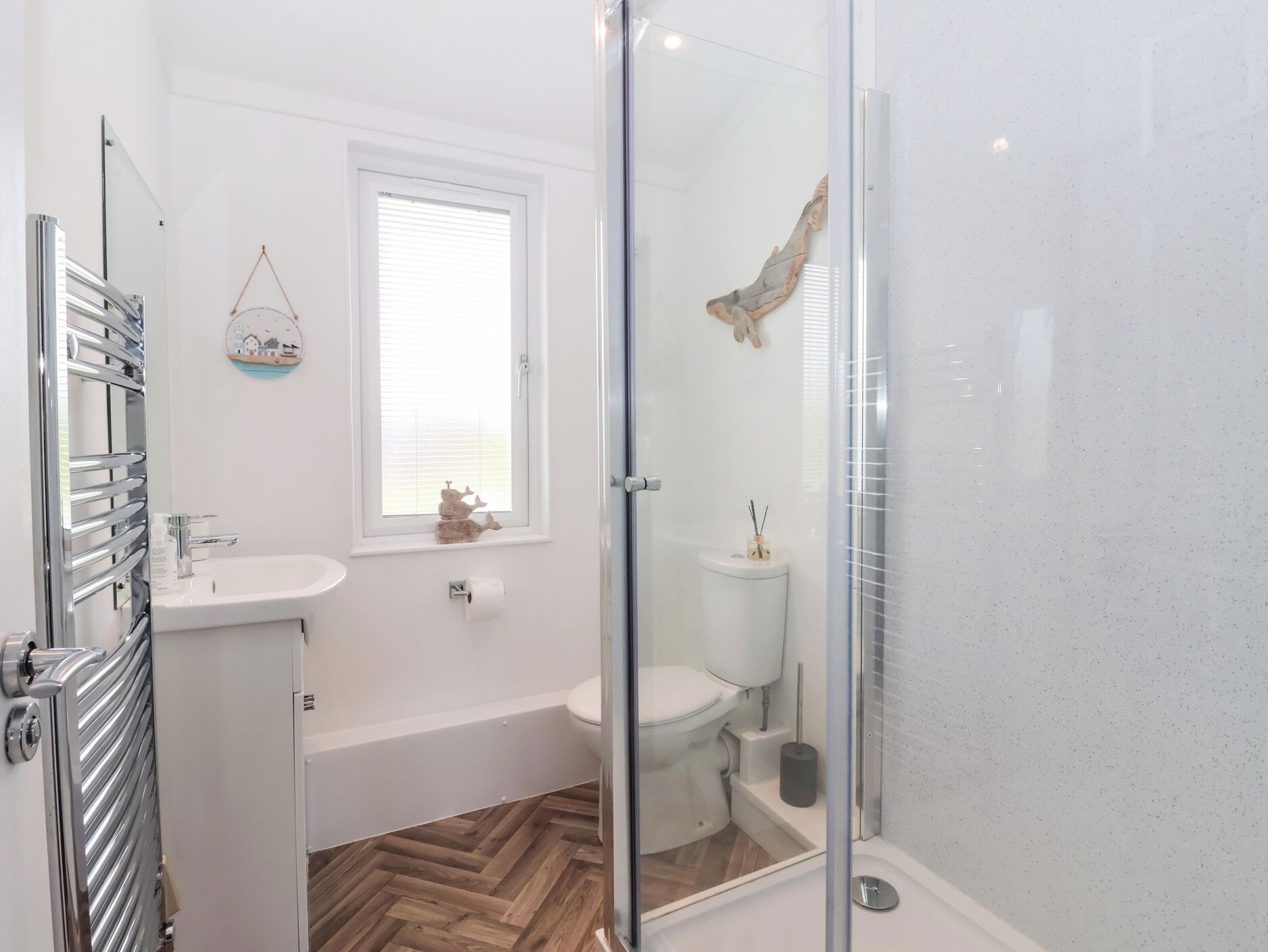 Un baño moderno con cabina de ducha de vidrio, un lavabo blanco, un inodoro y suelo de madera en patrón de espiga.