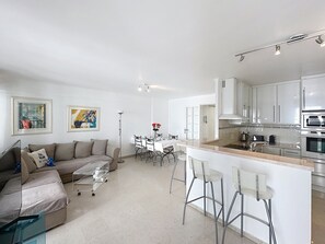 A modern kitchen with white cabinets, a central island with bar stools, a dining area with a white table and chairs, and a living area with a sofa and a coffee table.
