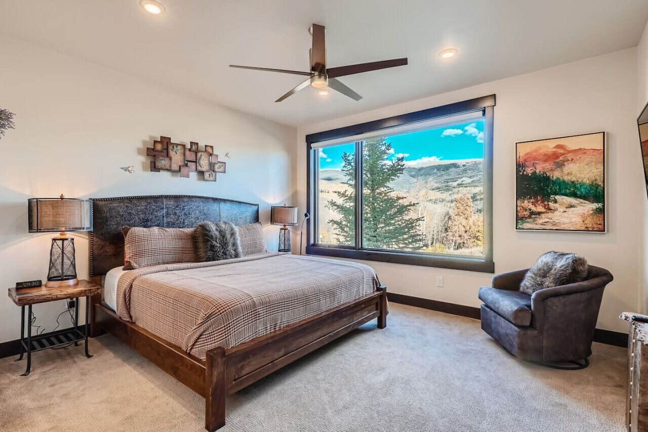 A bedroom with a large bed, a ceiling fan, a nightstand with a lamp, a chair, and a window with a mountain view.