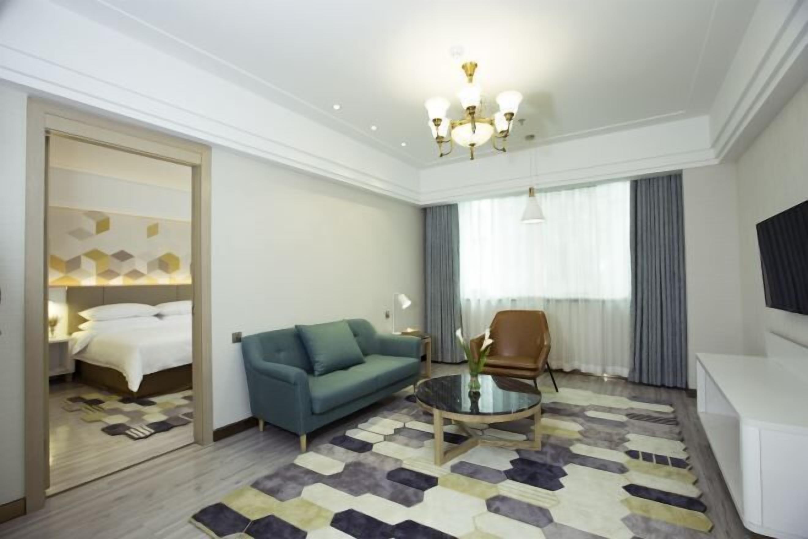 Habitación de hotel con cama, sofá azul, mesita de centro, televisor y un amplio ventanal con cortinas.