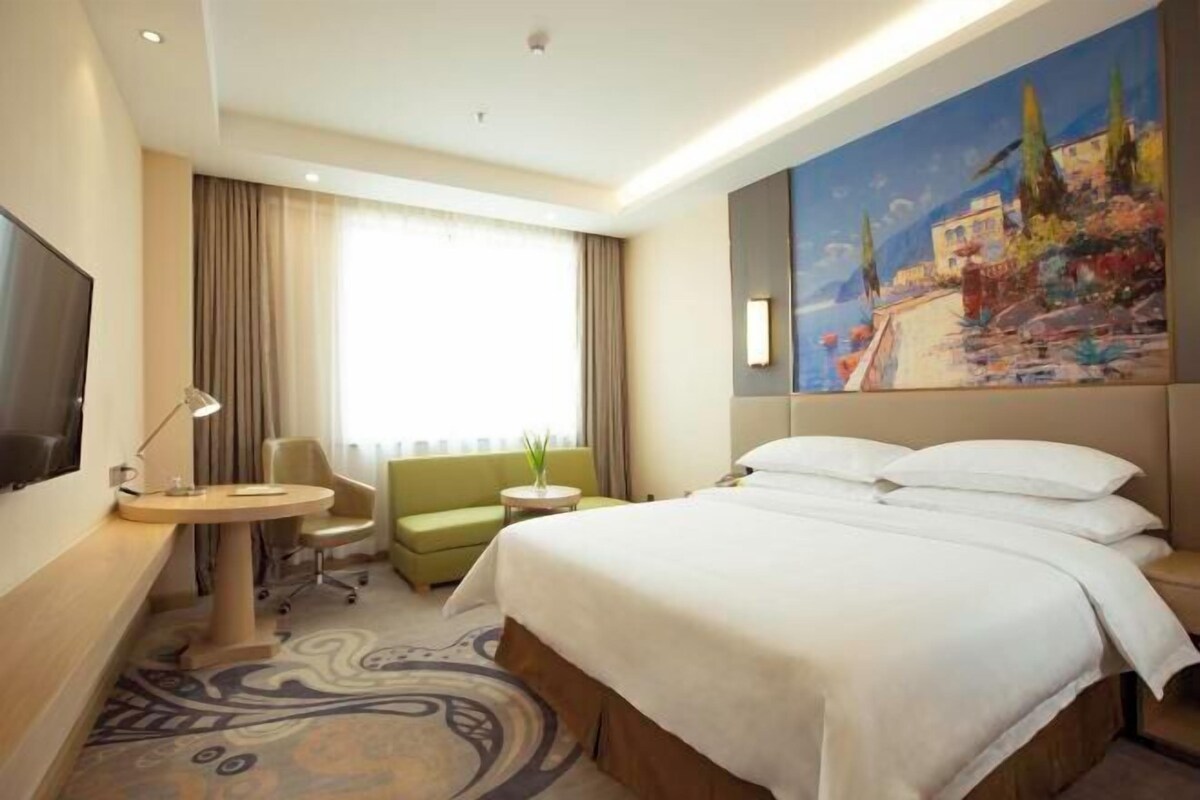 A hotel room with a bed, a desk, a chair, a small table, and a painting on the wall.