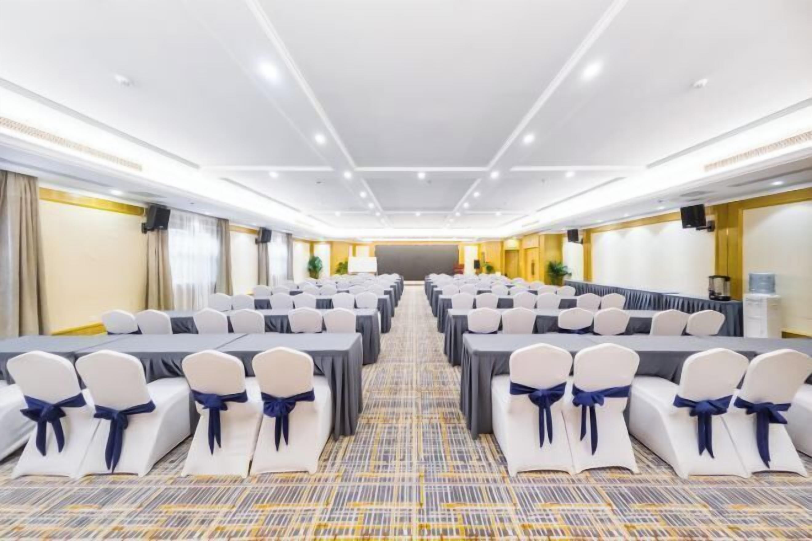 A conference room with rows of tables and chairs, a patterned carpet, and a white ceiling with recessed lighting.