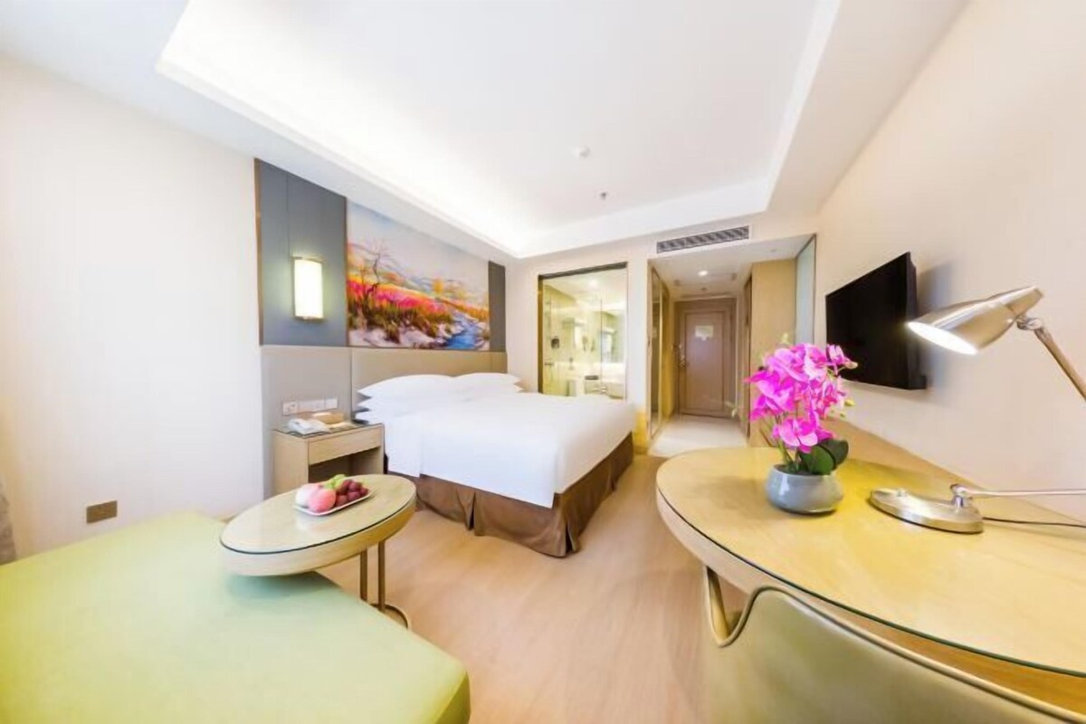 A hotel room with a bed, a desk, a chair, a small table, a TV, and a painting on the wall.