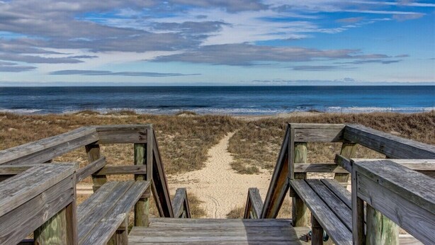 Un passeig de fusta que condueix a una platja, amb una vista clara de l'oceà i terres a la distància.