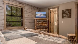 A bedroom with a large bed, a wooden headboard, a television on a wooden stand, a window with blinds, and a motivational quote on the wall.