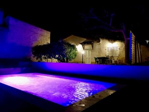 Ein Nachtblick auf einen Pool mit violetter Beleuchtung, eine Terrassenfläche mit Tisch und Stühlen sowie ein Gebäude mit einer Tür und Fenstern.