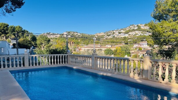 A rectangular pool with a balustrade, surrounded by a concrete deck. In the background, there are houses on a hillside under a clear blue sky.
