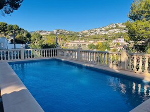 A rectangular pool with a balustrade, surrounded by a concrete deck. In the background, there are houses on a hillside under a clear blue sky. - 1.5km from L'Ampolla Beach and town | Comfortable | 3 double bedrooms | (Teulada)