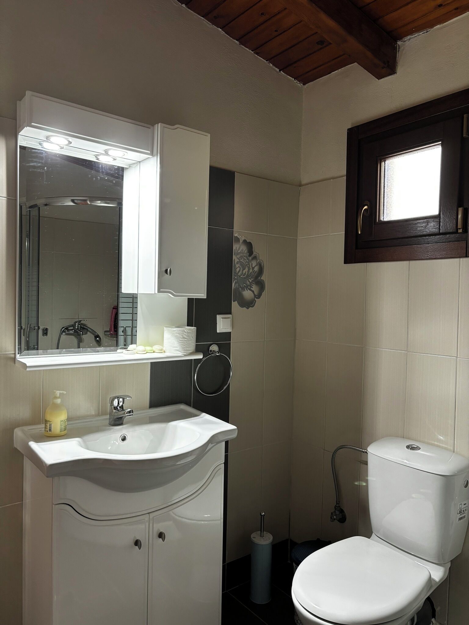 Un baño con lavamanos blanco, espejo, inodoro, zona de ducha y ventana sobre el inodoro.