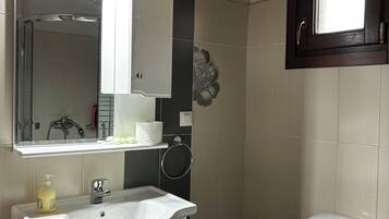 Un baño con lavamanos blanco, espejo, inodoro, zona de ducha y ventana sobre el inodoro.