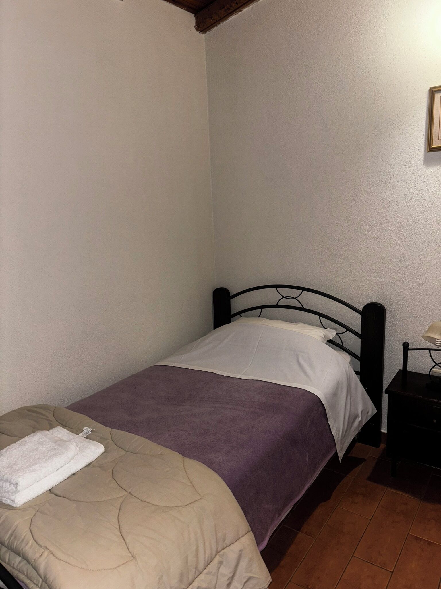 A small, single-bed room with a wooden headboard, a bedside table with a lamp, and a beige and purple bedspread.