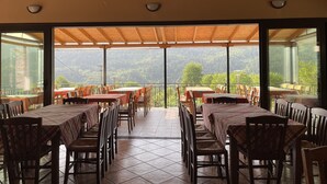 Een restaurant met een terras met uitzicht op een bergachtig landschap. Tafels staan onder een houten pergola, en de ruimte is afgesloten met glazen deuren.