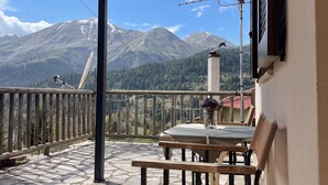 A wooden balcony with a stone floor, a table with chairs, and a view of snow-capped mountains.