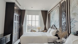 A hotel room with a large bed, a wardrobe, a desk, and a window with a view of the city. - Royal Hotel Phú Quốc (Phu Quoc)