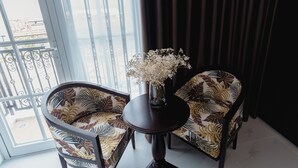 A room with two patterned armchairs, a small round table with a vase of flowers, and a view through a window with a balcony. - Royal Hotel Phú Quốc (Phu Quoc)