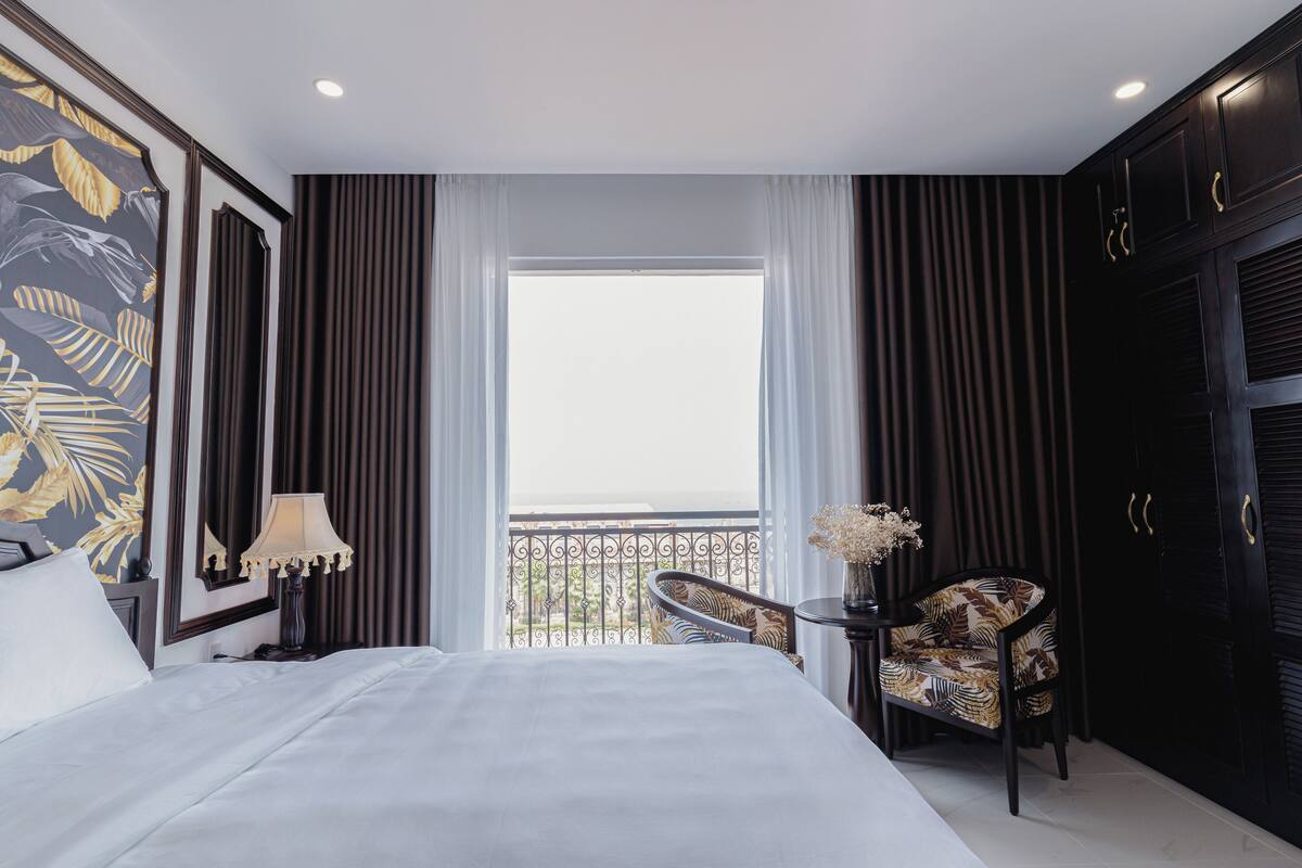 A hotel room with a large bed, a chair, a table with a vase, and a balcony with a railing.