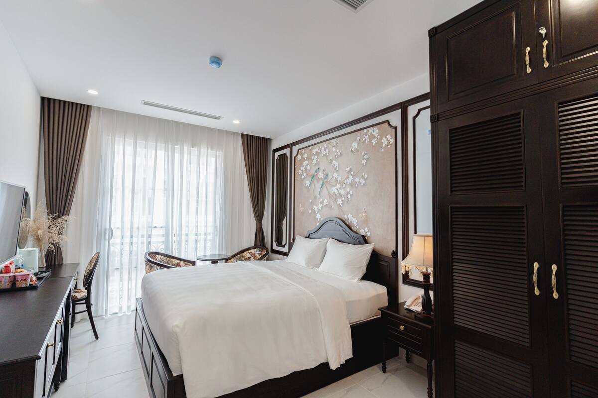 A hotel room with a large bed, a desk with a chair, a wardrobe, and a decorative wall panel.