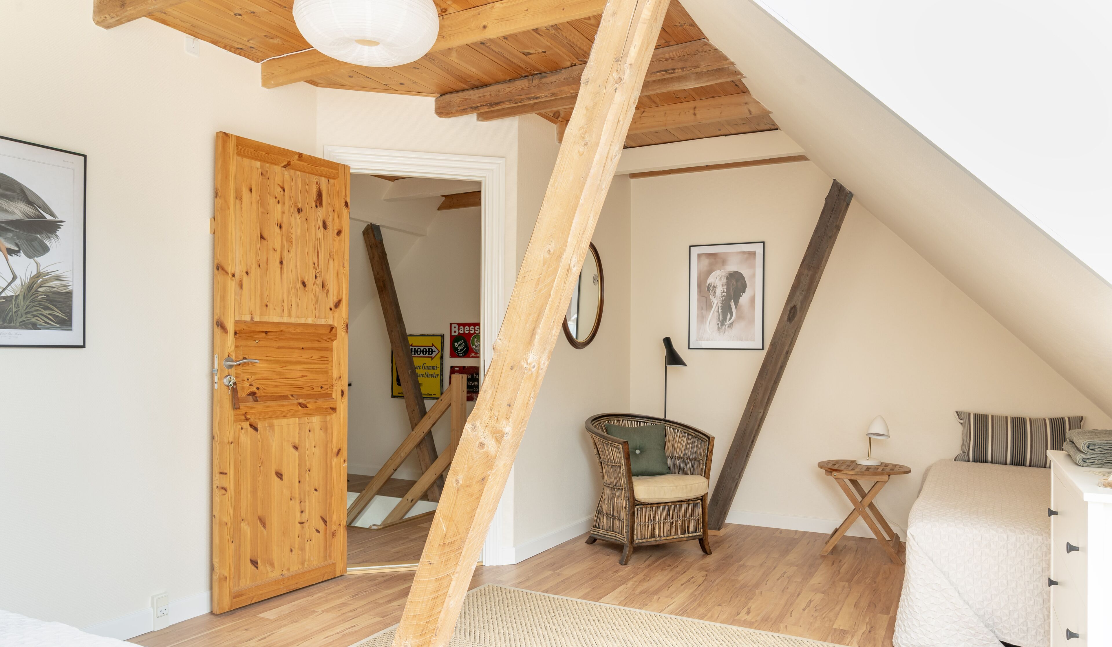 Una habitación con techo inclinado, vigas de madera, una silla de mimbre, una cama y una puerta que da a otra habitación.