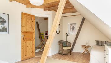 Una habitación con techo inclinado, vigas de madera, una silla de mimbre, una cama y una puerta que da a otra habitación.