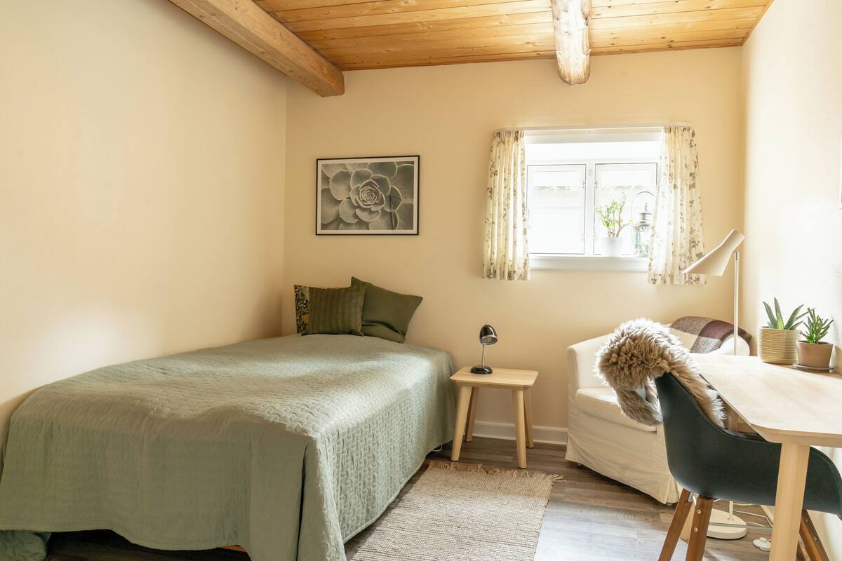 A bedroom with a bed, a small table with a lamp, a chair, a window with curtains, and a potted plant.