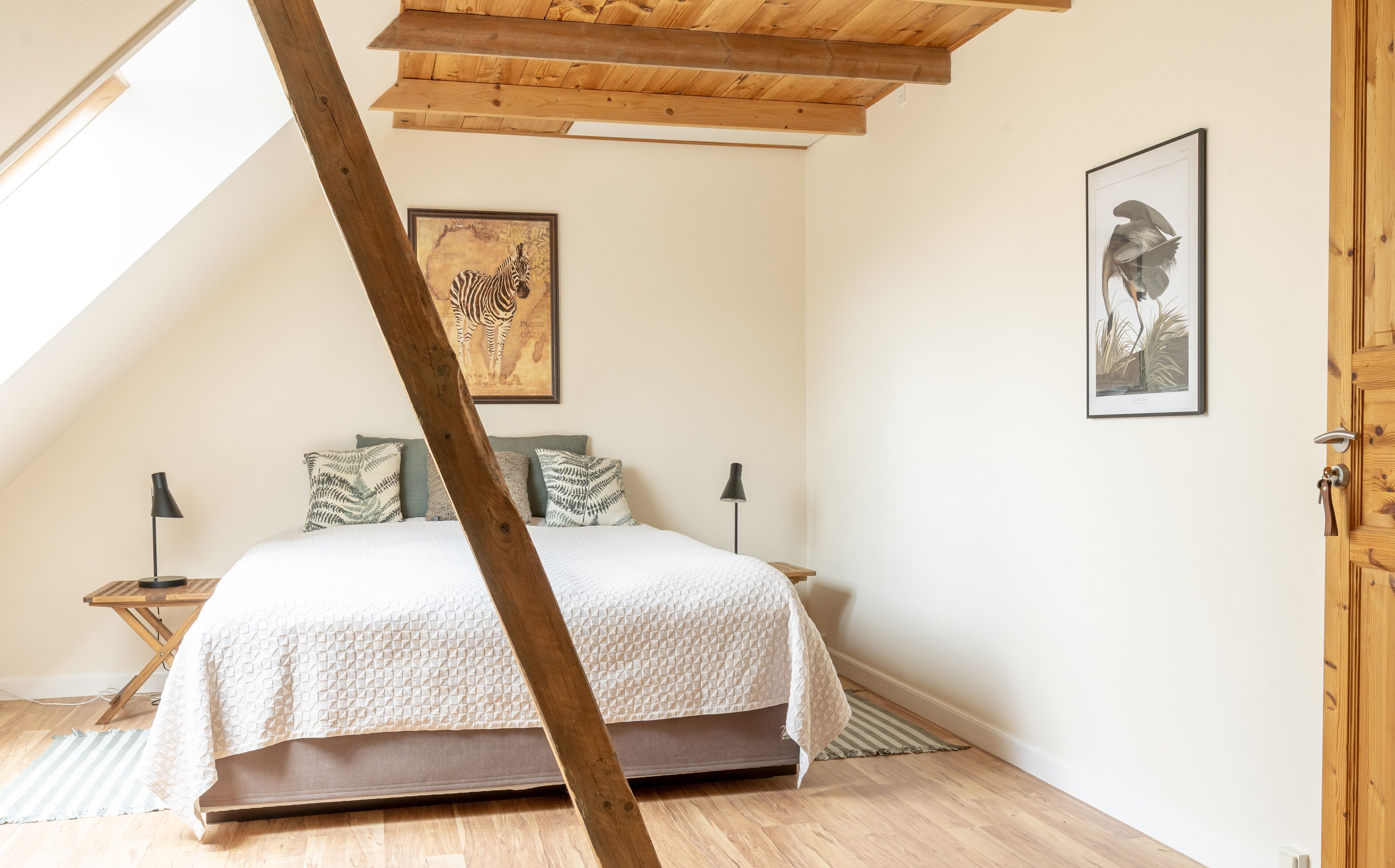 A bedroom with a sloped ceiling, a bed with a white quilt, two nightstands, a framed zebra picture, and a framed plant illustration.