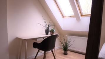 A room with a sloped ceiling, a desk with a chair, two potted plants, and a radiator.
