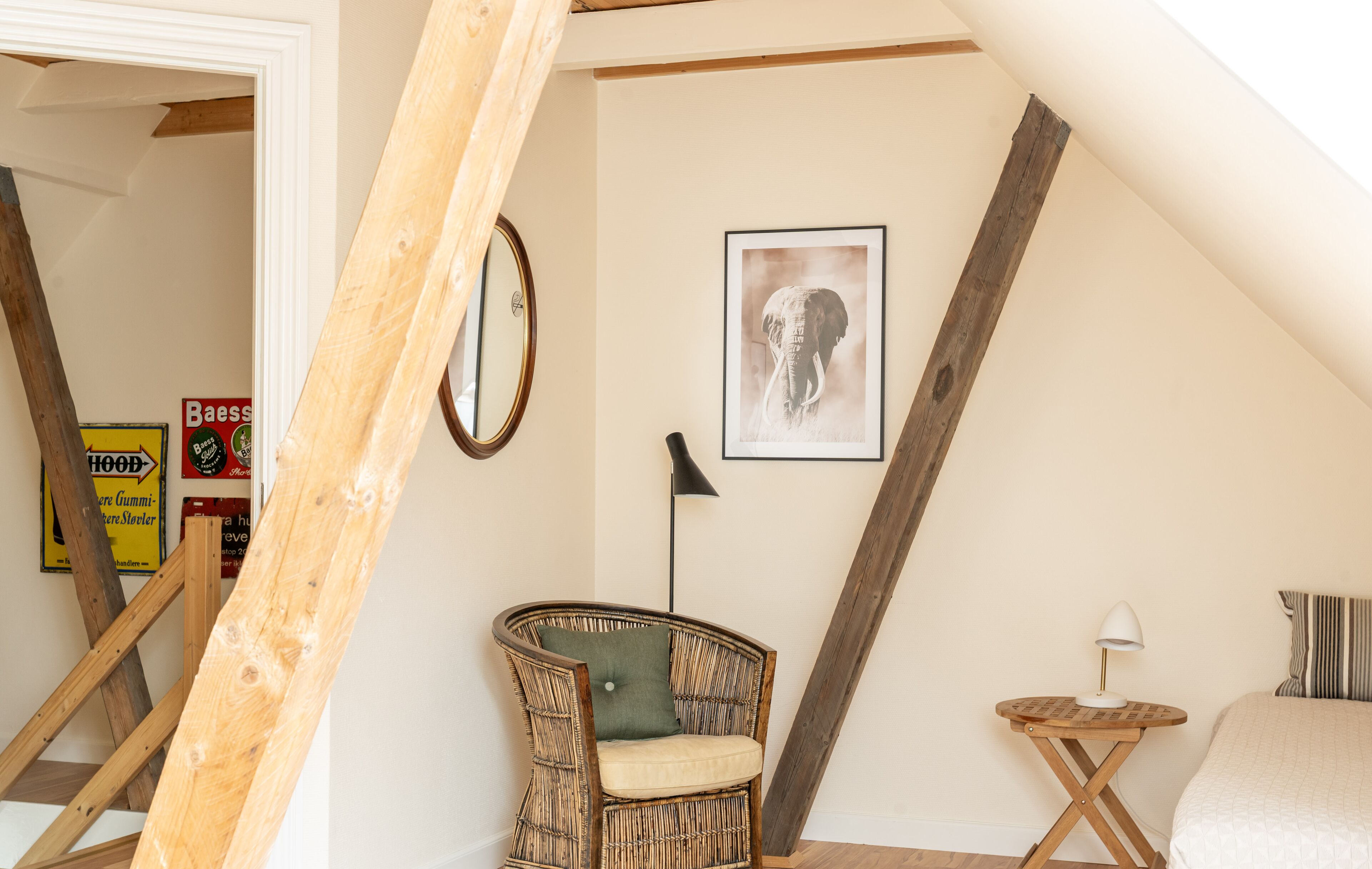 Une pièce avec un plafond haut, une chaise en osier avec un coussin, un tableau représentant un éléphant encadré, une petite table ronde avec une lampe, et un lit avec des oreillers à rayures.