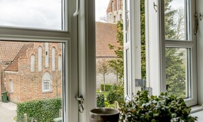 A view through a window of a brick building with a tiled roof, a stone wall, and a tree-lined garden. - Akaciegaarden Bed & Breakfast (Harlev)
