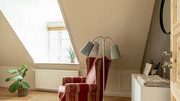 Una habitación acogedora con techo inclinado, un sillón tapizado de rojo, una lámpara de pie, un radiador, una planta en maceta y un aparador de madera con espejo.