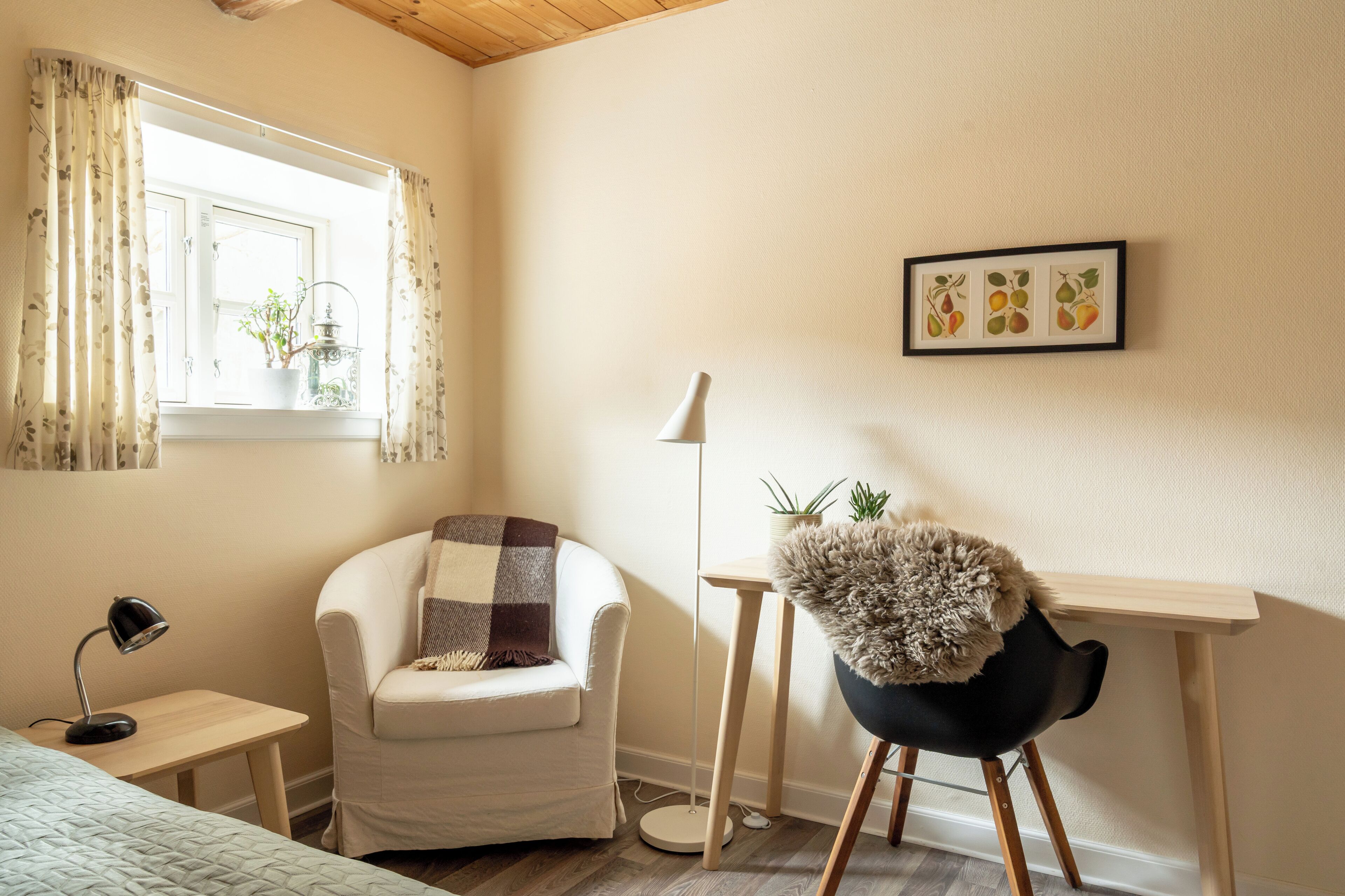 A cozy room with a white armchair, a desk with a lamp, a small table with a lamp, and a window with curtains.