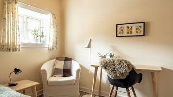 A cozy room with a white armchair, a desk with a lamp, a small table with a lamp, and a window with curtains.