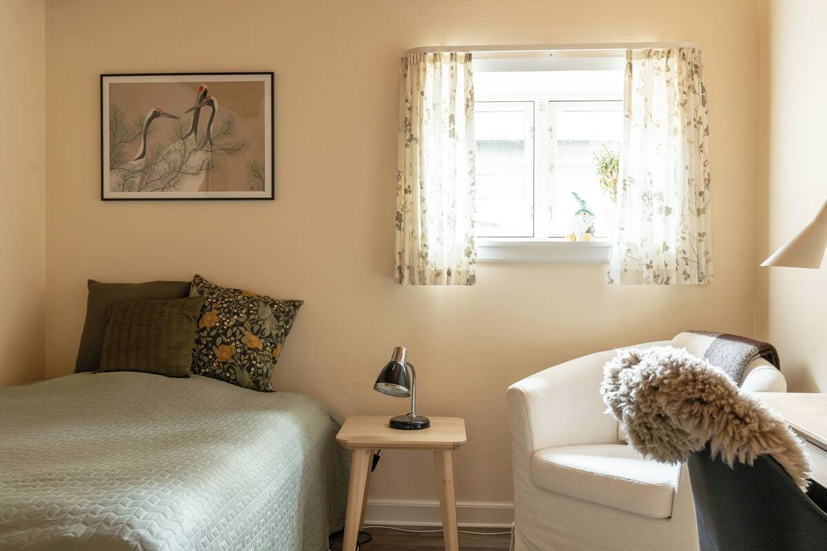 A bedroom with a bed, a small bedside table with a lamp, a chair, a window with curtains, and a wall-mounted picture.